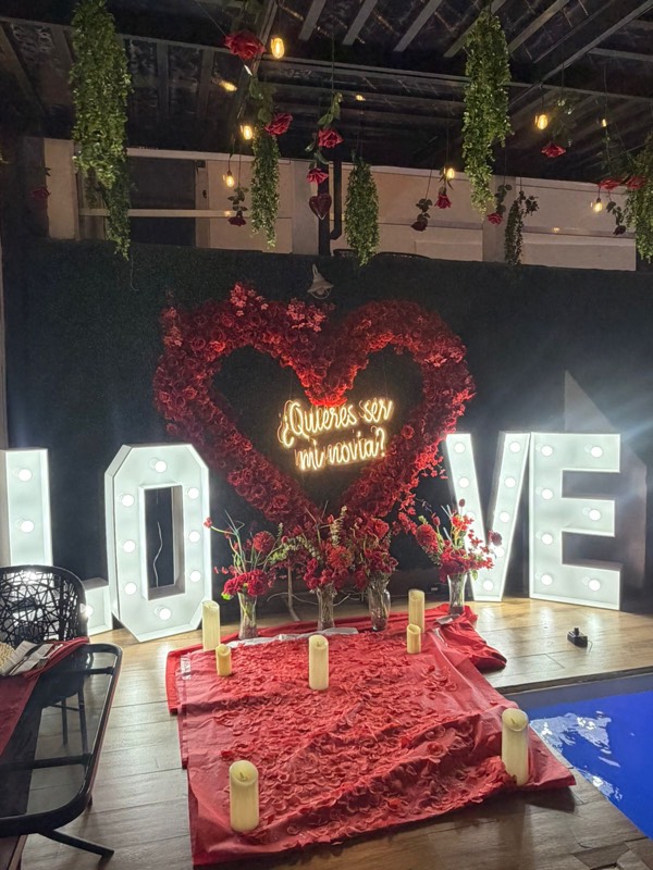 Letras LOVE iluminadas y corazón de rosas rojas con letrero de neón «¿Quieres ser mi novia?», velas y flores, junto a la alberca