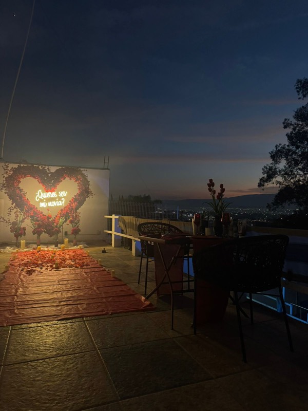 Corazón de rosas con neón en la terraza al anochecer, mesa montada y la ciudad iluminada al fondo